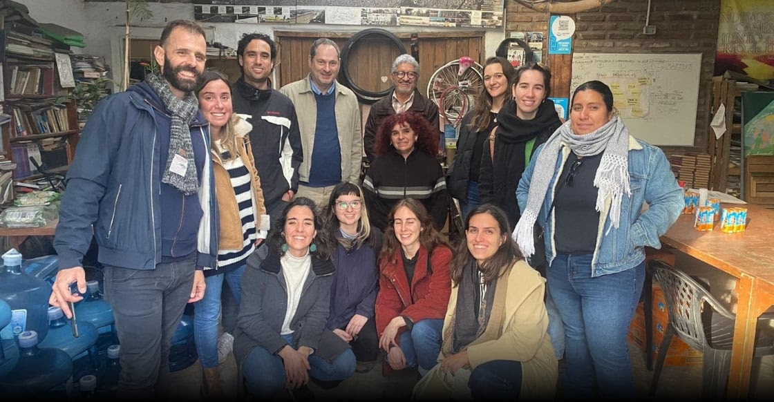 Member representatives of the Latin America PAR projects and the Grassroots Justice Network standing and kneeling together smiling at the camera. They are inside a room with a bookshelf and wooden table, a piece of paper hangs in the background with a map of their notes.