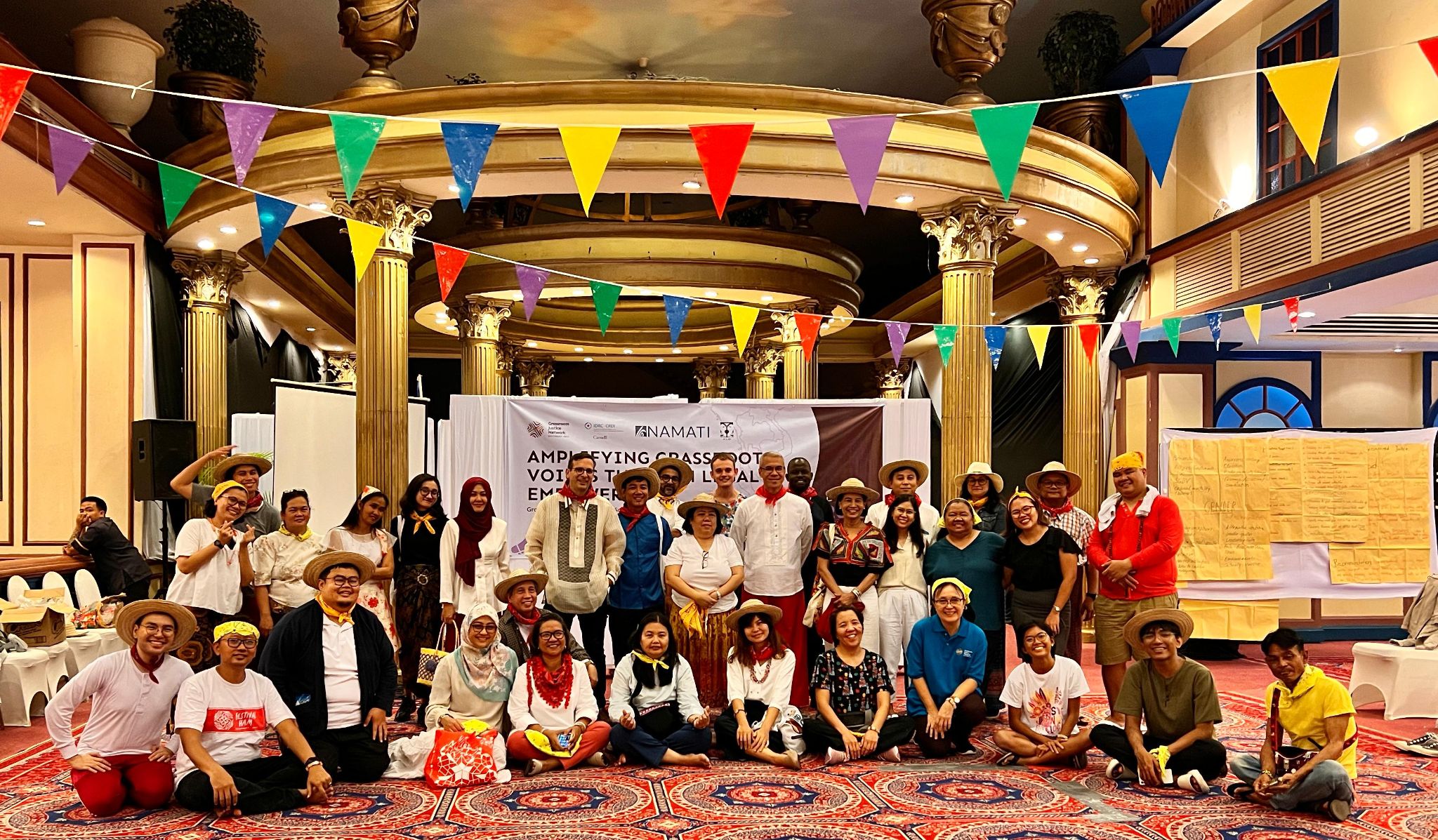  A large group of participants from the convening face the camera and smile, sit inside a large hall decorated with colorful banners and a Namati banner behind them.