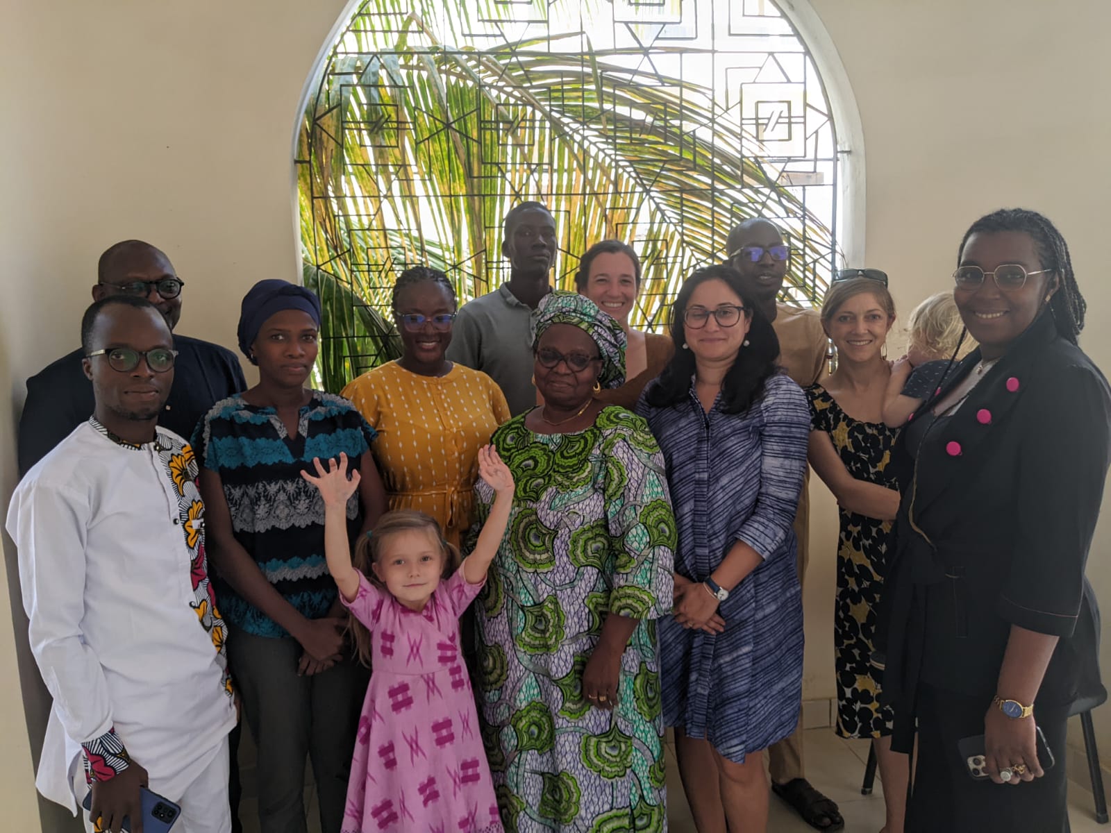 Participating members of the West Africa convening in Dakar stand in a group and face the camera smiling. They are standing in front of a window with a decorative screen of geometric shapes, and a palm tree outside the window.