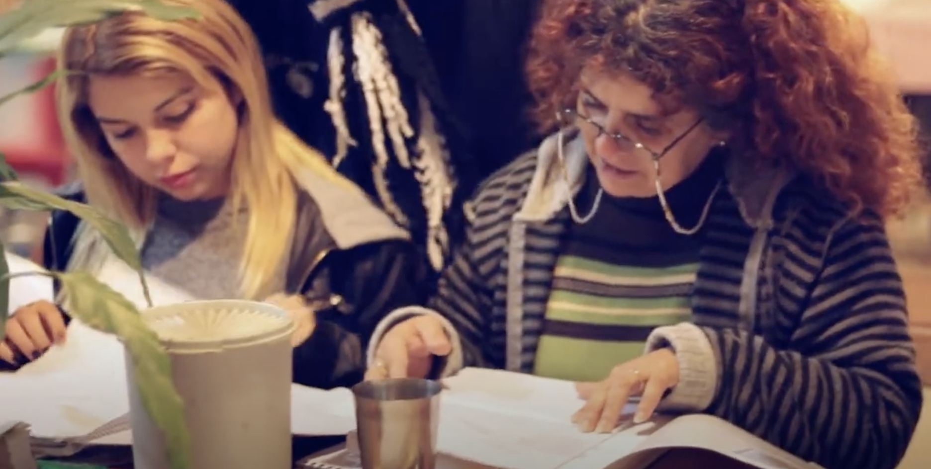 Two women sit at a table pouring over documentation. On the left, a woman with straight blond hair, and on the right, a woman with red-brown curly hair and wearing reading glasses.