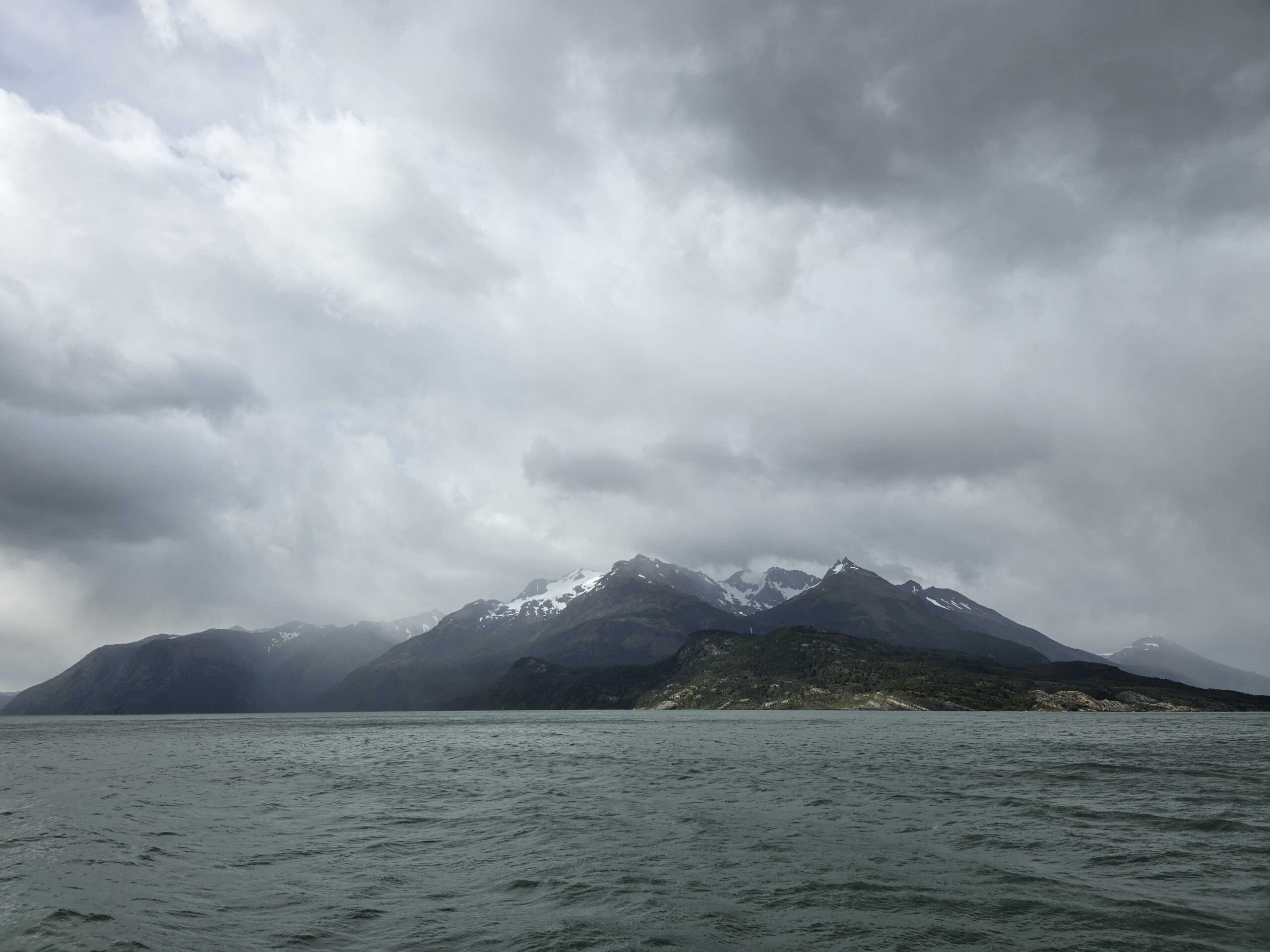 A snow-capped mountain rises above a body of water under a cloudy sky.
