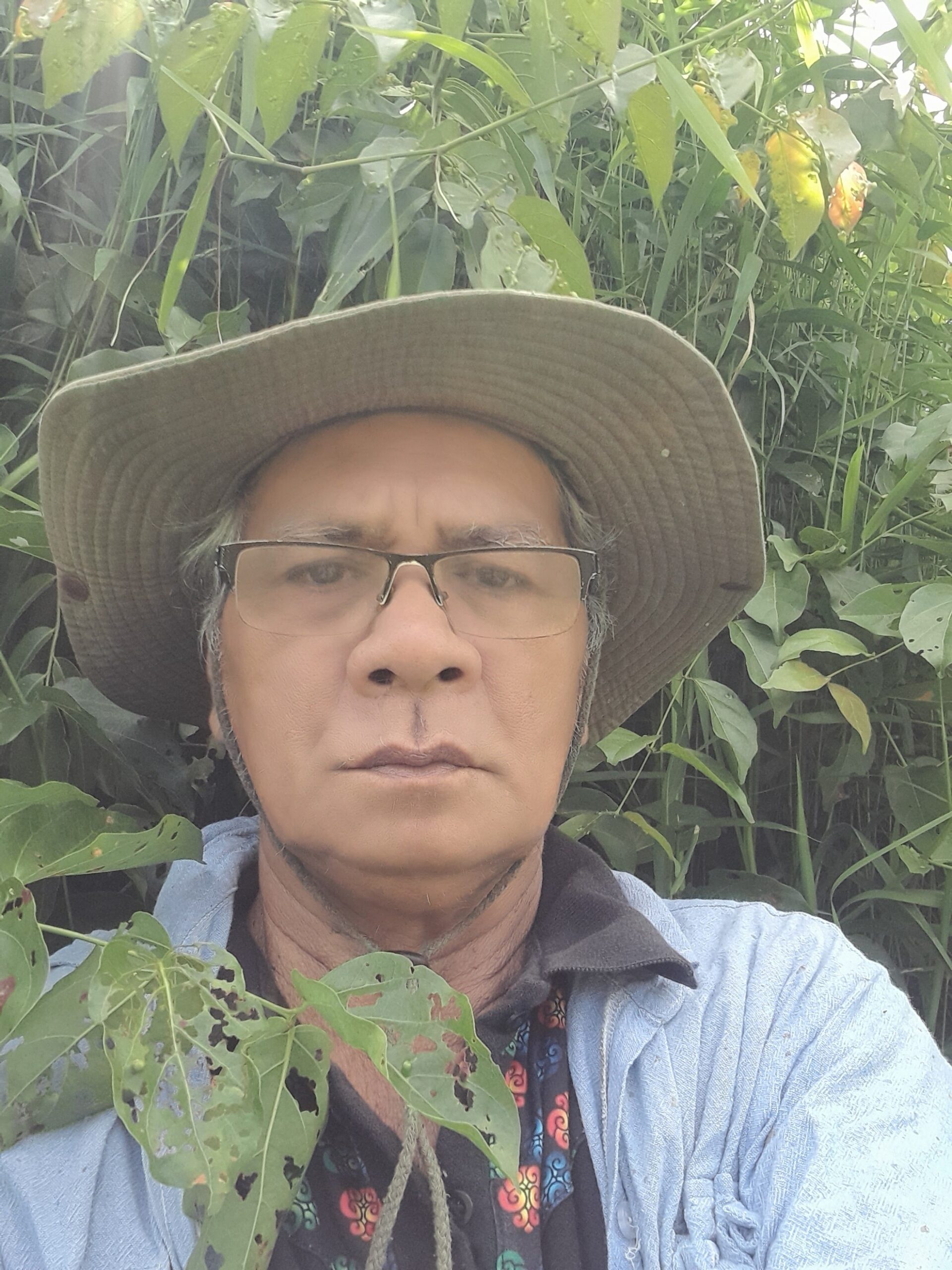 A man in a hat takes a serious selfie in front of green foliage.