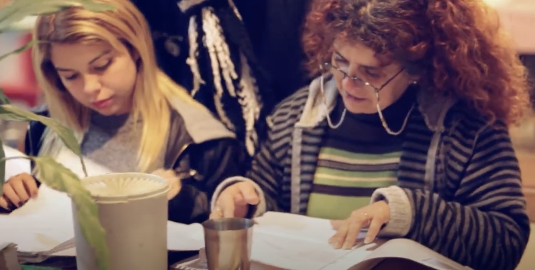 Dos mujeres están sentadas a una mesa revisando unos documentos. A la izquierda, una mujer con el pelo liso y rubio; a la derecha, una mujer con el pelo rizado de color castaño rojizo y gafas de lectura.