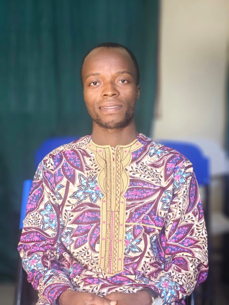 A man in a colorful purple traditional shirt sits and smiles at the camera.