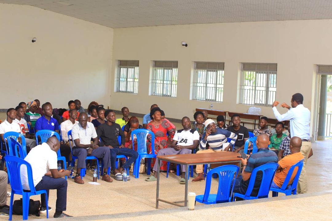 A man speaks to a group of adults in blue chairs in a large room.