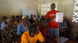 Man in red shirt showing a certificate to a group of seated men in a classroom. Whiteboard and lattice window in background.